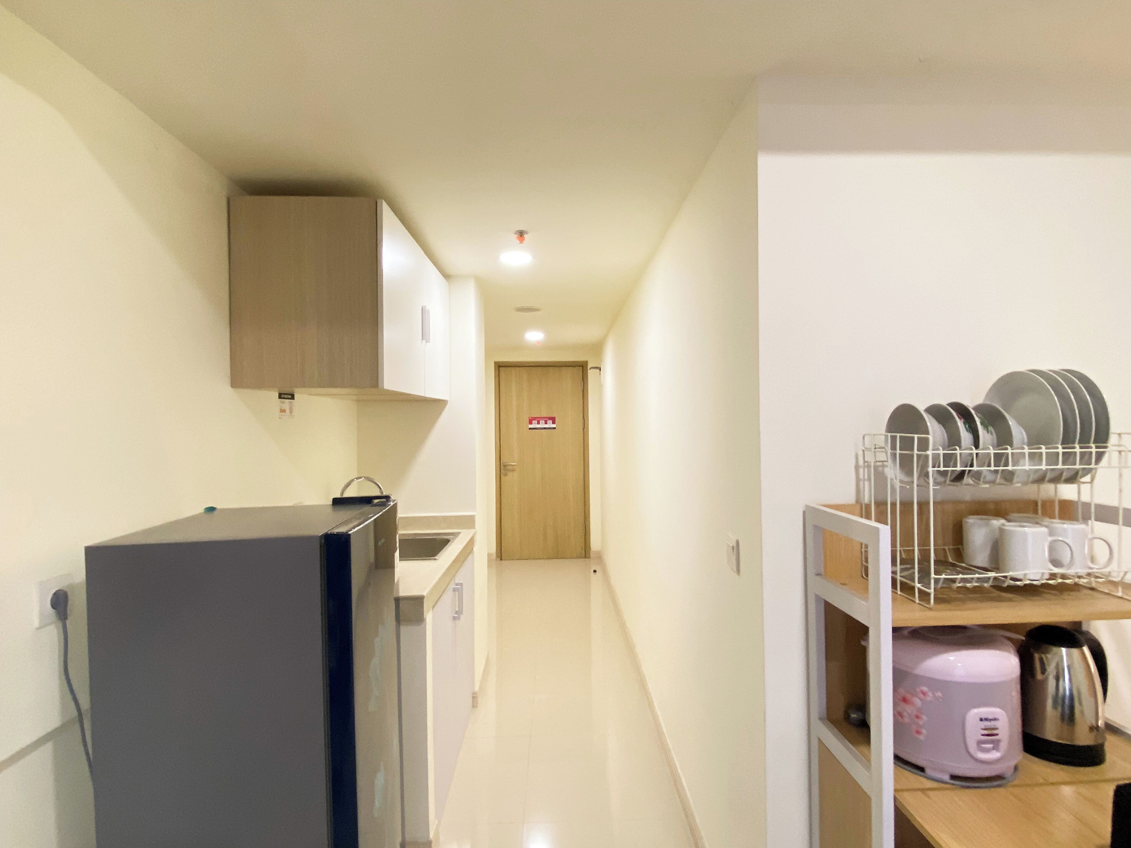 A narrow hallway with a refrigerator, a microwave, and a sink on the left. A door with a red sign is visible in the distance.