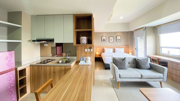 A modern hotel room with a kitchenette, a bed with two pillows, a grey sofa, and a window with a view. - Spacious And Elegant Studio Apartment Mustika Golf Residence (Cikarang)