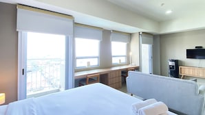 A hotel room with a bed, a desk, a chair, a TV, and a window with a view of the city. - Spacious And Elegant Studio Apartment Mustika Golf Residence (Cikarang)