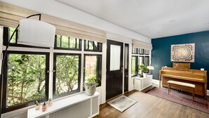 Une pièce avec un piano, un radiateur, un banc et une grande fenêtre avec des stores.