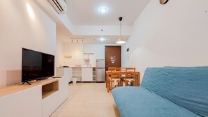 A modern living room with a flat-screen TV, a blue sofa, wooden chairs, a kitchen area with a fridge, and a dining table.