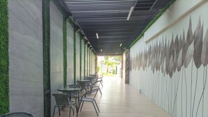 Un pasillo con sillas y mesas de mimbre, detalles verdes en la pared y un mural con motivos de hojas.