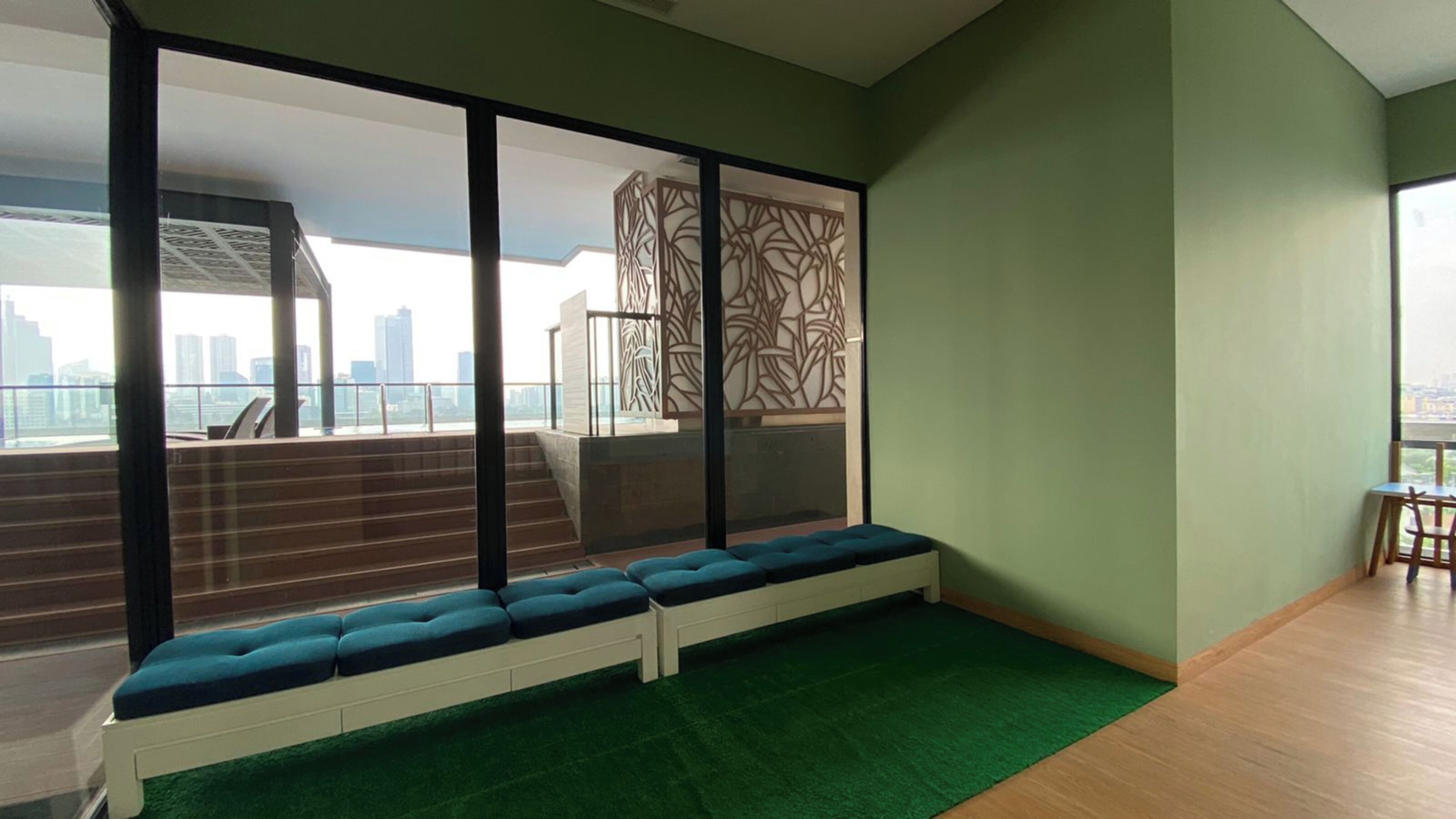 A modern room with a green carpet, a white bench with blue cushions, a large window with a city view, and a patterned wall.