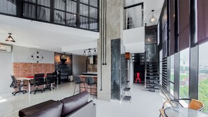 A spacious living area with a high ceiling, a dining area with modern furniture, a kitchen with a brick accent wall, and large windows offering a view of the city. - Spacious 2Br Loft At Brooklyn Alam Sutera Apartment (South Tangerang)
