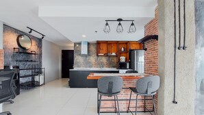 A modern kitchen with a brick backsplash, wooden cabinets, and a central island. There are two bar stools and a round table with a black chair. - Spacious 2Br Loft At Brooklyn Alam Sutera Apartment (South Tangerang)