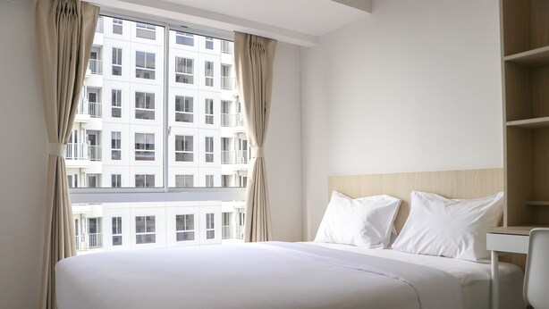 A hotel room with a large bed, a wooden headboard, and a view of a building through a window with curtains. - Restful And Cozy 2Br Tokyo Riverside Pik 2 Apartment (Teluknaga)