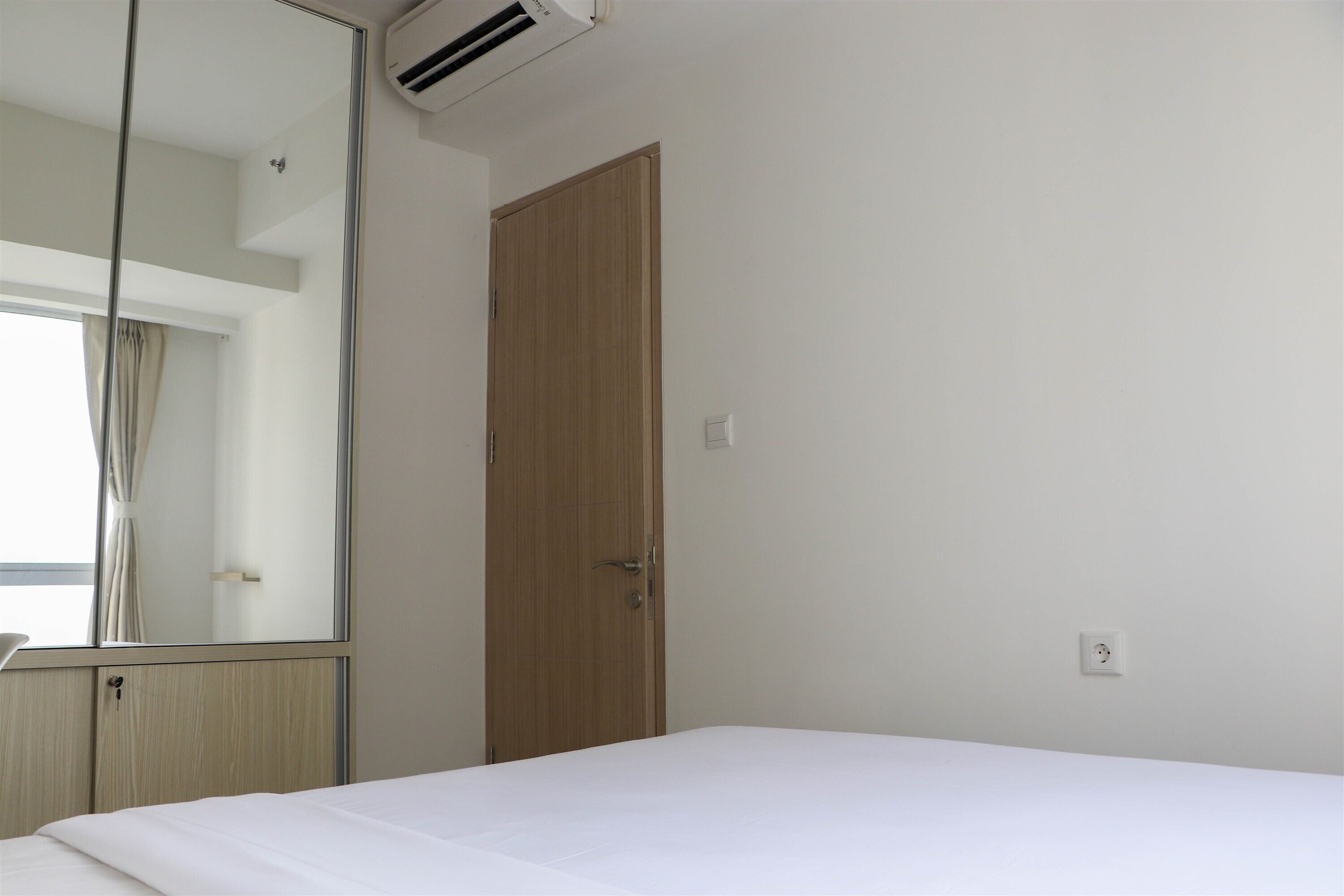 A hotel room with a wooden door, a bed, a mirror, and an air conditioning unit.