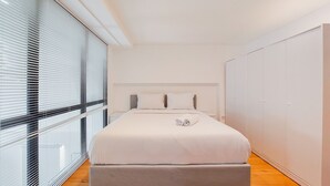 A modern bedroom with a large bed, a grey bench, and a wardrobe. - Spacious And Elegant 3Br Loft At Kingland Avenue Apartment (South Tangerang)