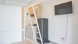 A compact kitchen with a refrigerator, a TV mounted on the wall, and a white ladder leaning against the cabinets. - Simply Look And Homey Studio Tokyo Riverside Pik 2 Apartment (Teluknaga)