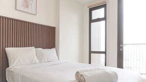 A neatly made bed with white linens and a rolled towel, a wooden headboard, and a large window with a view. - Brand New And Comfort Studio At Vasaka Solterra Apartment (Jakarta)