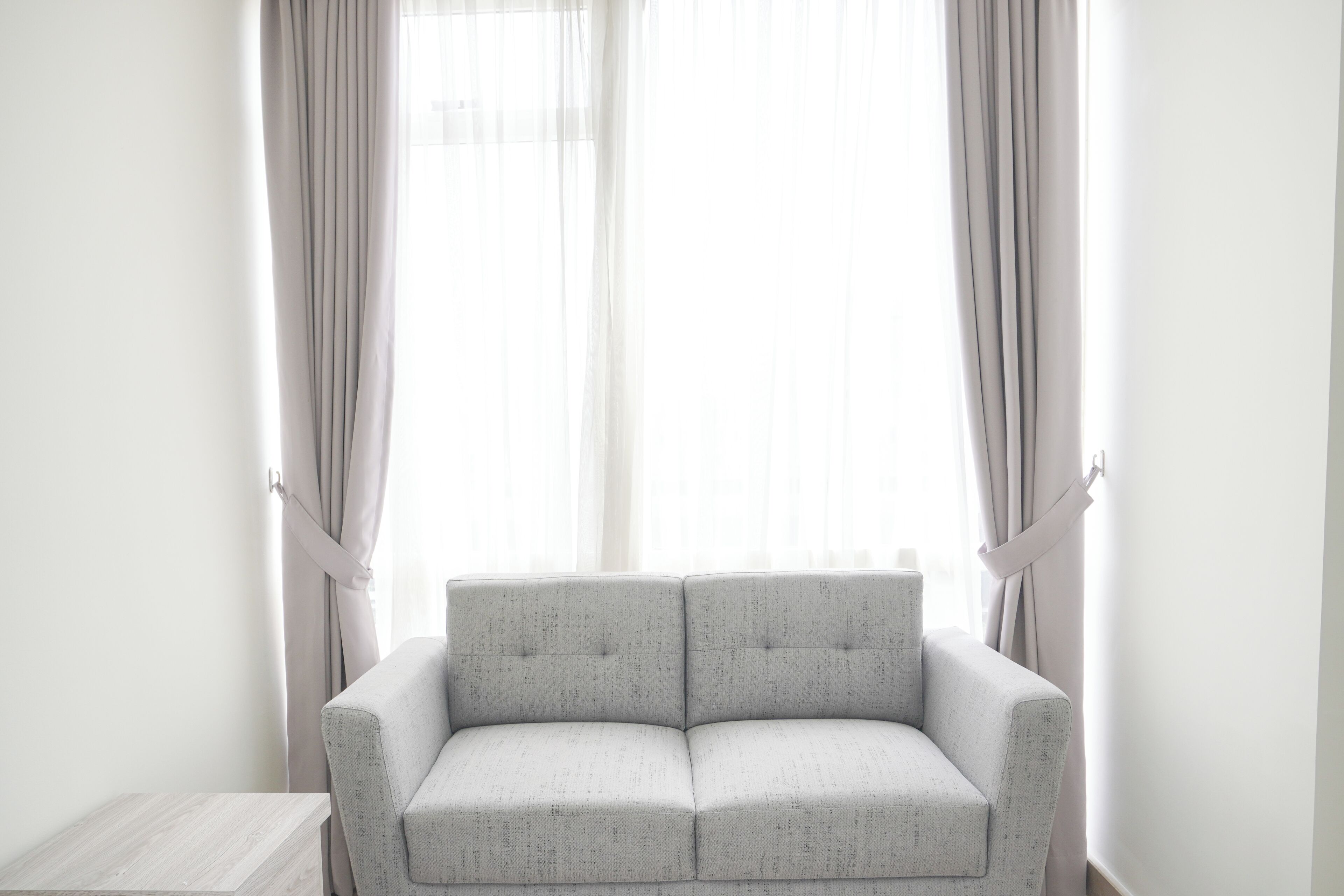 Una habitación minimalista con un sofá gris, una pequeña mesita de madera y cortinas translúcidas en una ventana.