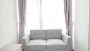 Et minimalistisk rum med en grå sofa, et lille træsidebord og lette gardiner foran et vindue.