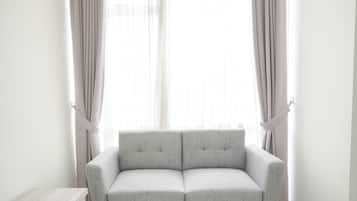 Una habitación minimalista con un sofá gris, una pequeña mesita de madera y cortinas translúcidas en una ventana.