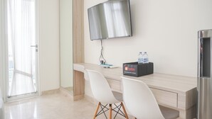 Et moderne hotelværelse med et træskrivebord, to hvide stole, et fladskærms-tv monteret på væggen, en minibar og en dør med let gardin.