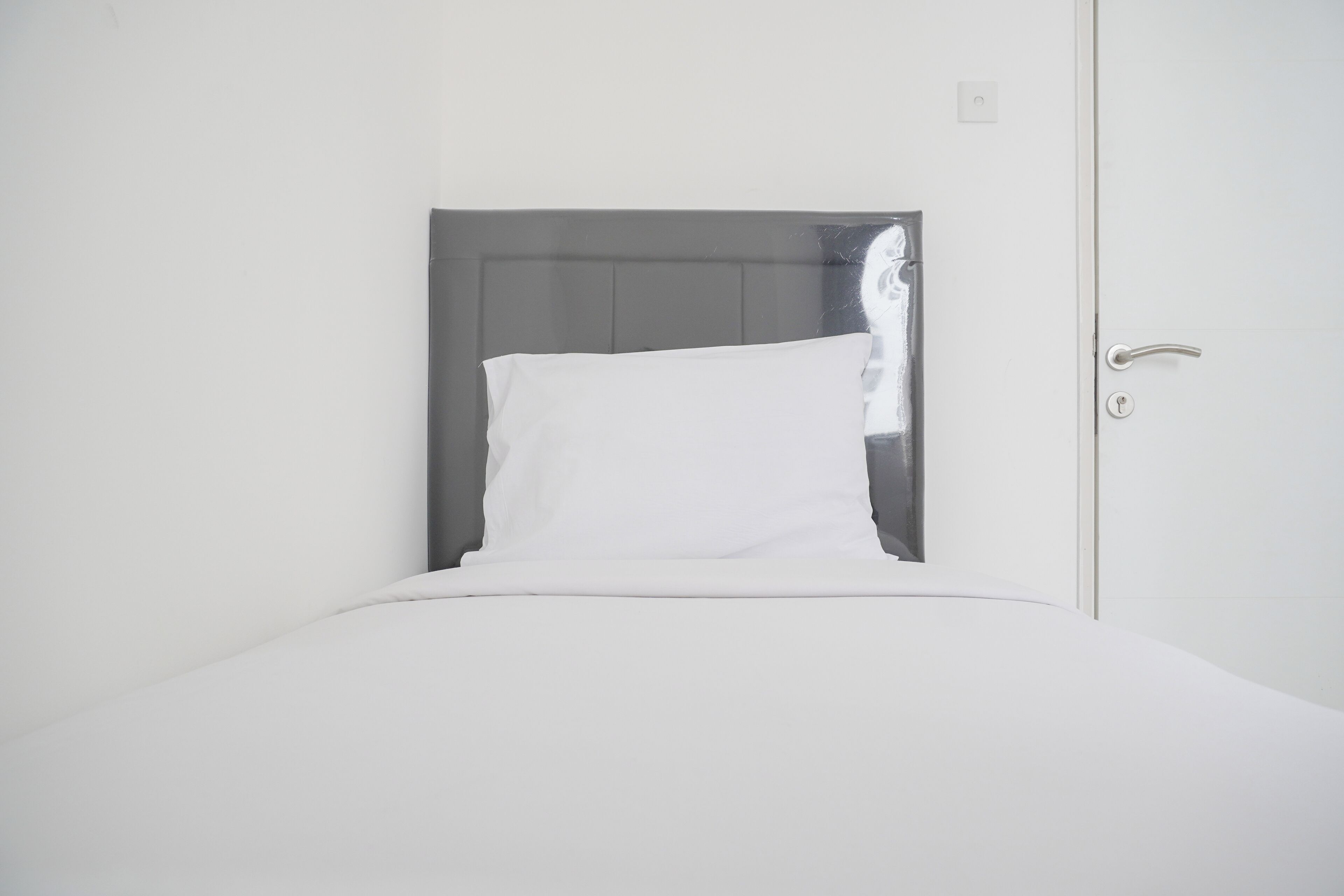 Una cama bien hecha con ropa blanca y un cabecero gris en una habitación con paredes blancas y una puerta.