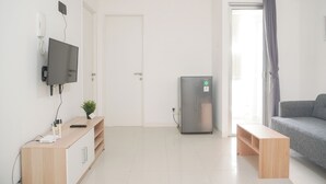 A modern living room with a flat-screen TV mounted on the wall, a built-in refrigerator, a grey sofa, and a wooden bench. - Best Deal And Comfort 3Br At Bassura City Apartment (Jakarta)