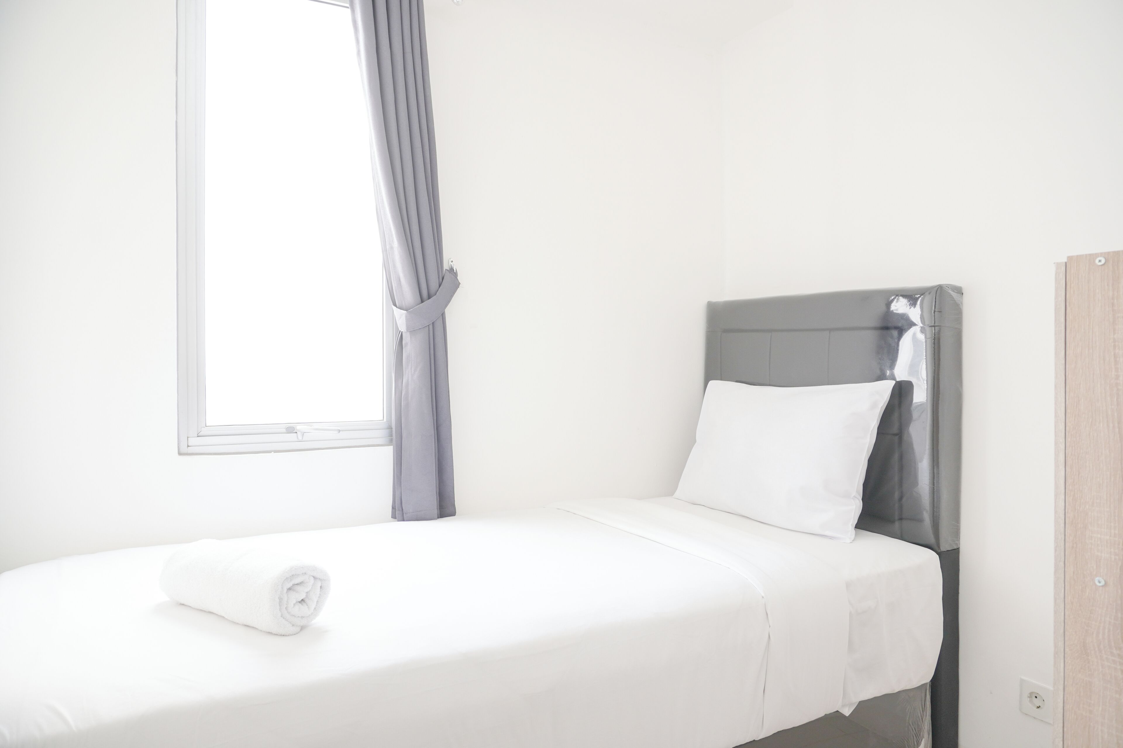 A single bed with a white mattress and grey headboard in a bright room with a window and curtains.