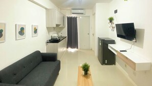 A modern living room with a grey sofa, a wooden coffee table, a flat-screen TV, and a kitchen area with a refrigerator and a small plant. - Best Deal And Homey 2Br Bassura City Apartment (Jakarta)