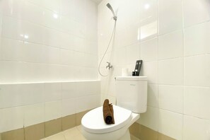 A white toilet with a brown towel on it, a white toilet paper holder, and a shower head on the wall. - Comfort Living And Strategic 2Br At Bassura City Apartment (Jakarta)
