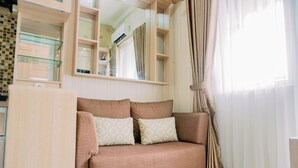 A living room with a beige sofa, patterned cushions, a glass shelf unit, and a window with sheer curtains. - Comfort Stay 2Br At Green Pramuka City Apartment (Jakarta)