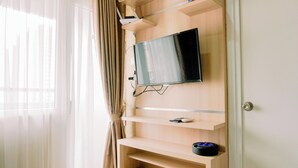 A room with a mounted TV on a wooden wall, a window with sheer curtains, and a door with a silver handle. - Comfort Stay 2Br At Green Pramuka City Apartment (Jakarta)