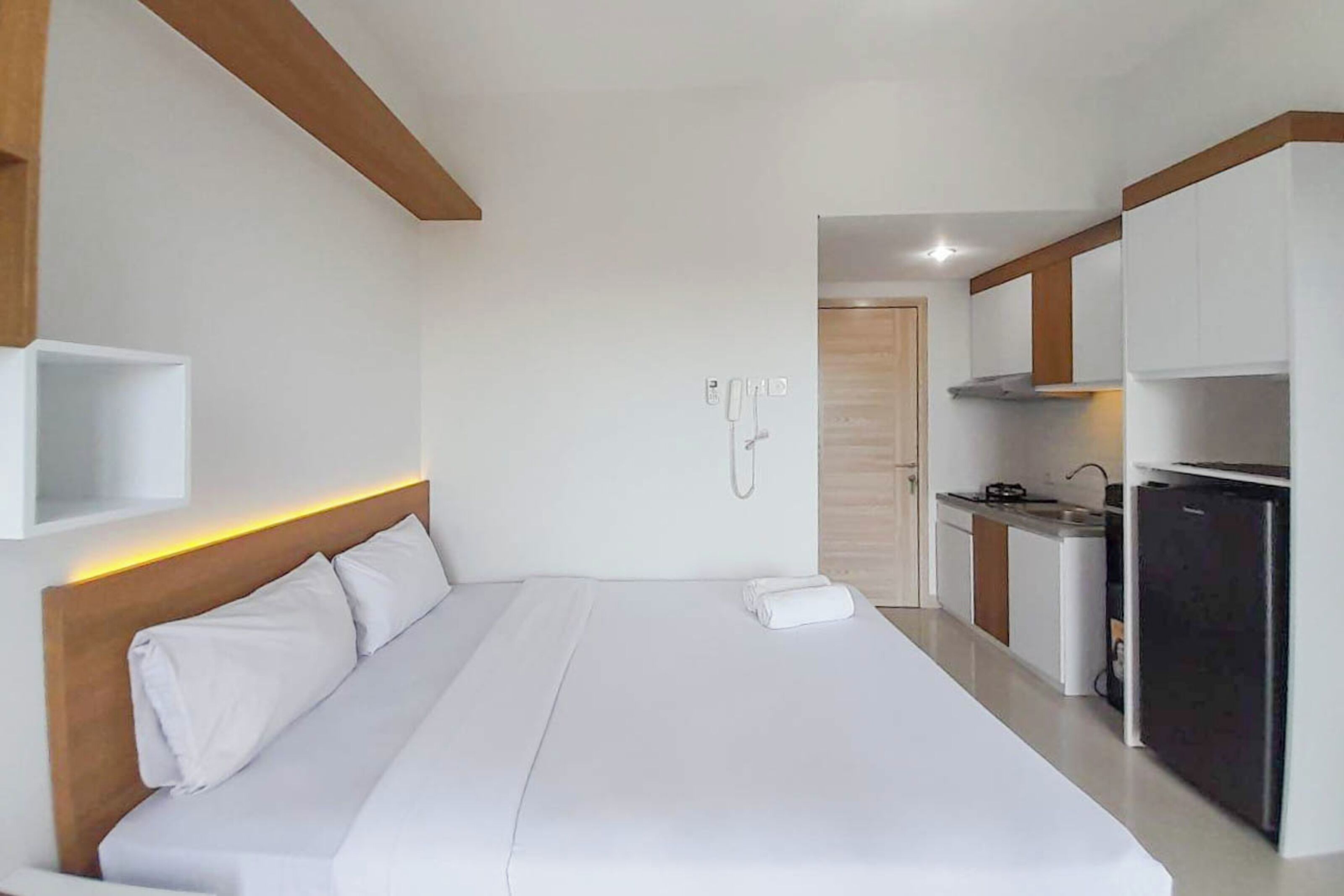 A modern hotel room with a large bed, a small kitchenette, and a wooden ceiling beam.