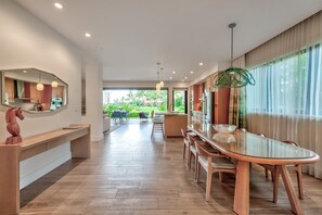 A modern hotel lobby with a long reception desk, a glass-top dining table with chairs, and a view of a lush garden through large windows.