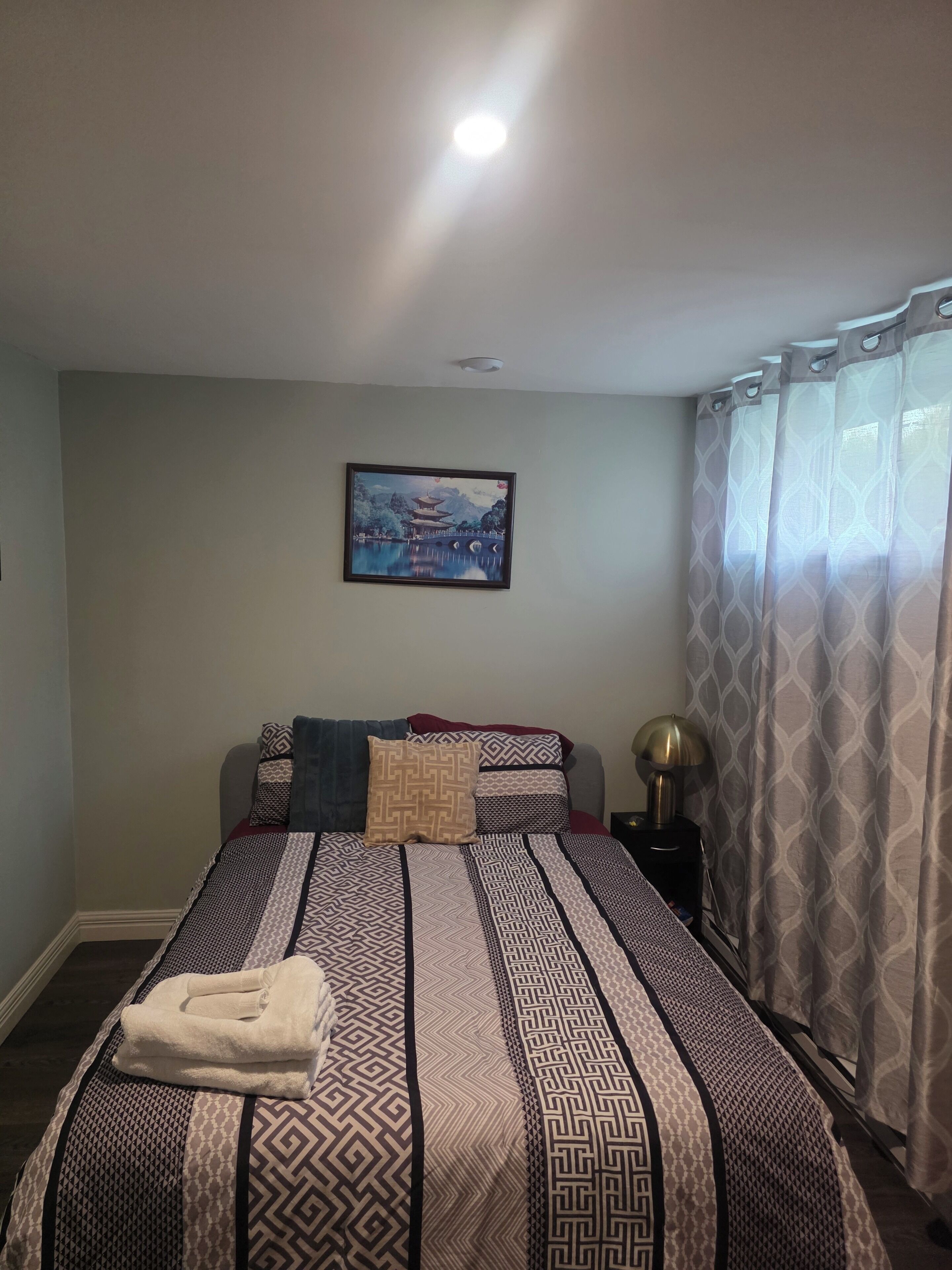 Un dormitorio con cama, almohadas, una colcha, un cuadro en la pared, una lámpara y cortinas.