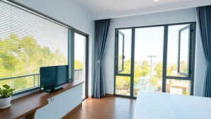 Een slaapkamer met een bed, een televisie op een houten plank, grote ramen en uitzicht op groen buiten.
