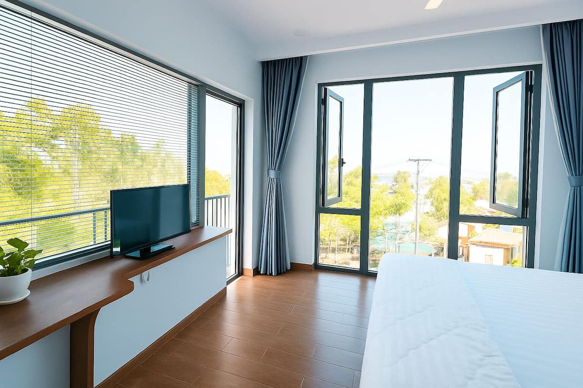 Une chambre à coucher avec un lit, un téléviseur posé sur une étagère en bois, de grandes fenêtres et une vue sur la verdure à l’extérieur.