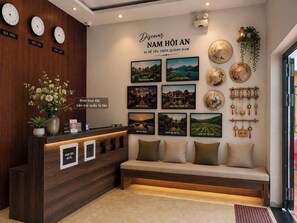 Un espace d’accueil d’hôtel comprenant un comptoir de réception en bois, un banc avec des coussins et un mur décoré d’horloges et de tableaux représentant des paysages.