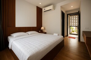 Quarto de hotel com uma cama grande, painéis de madeira, um aparelho de ar condicionado, uma mesa de cabeceira com abajur e vista através de uma porta.
