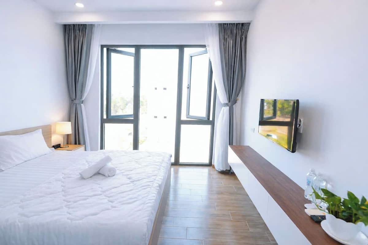 Une chambre d’hôtel avec un lit, un téléviseur fixé au mur, une table de chevet en bois ornée d’une plante, et une vue sur l’extérieur par la porte ouverte.