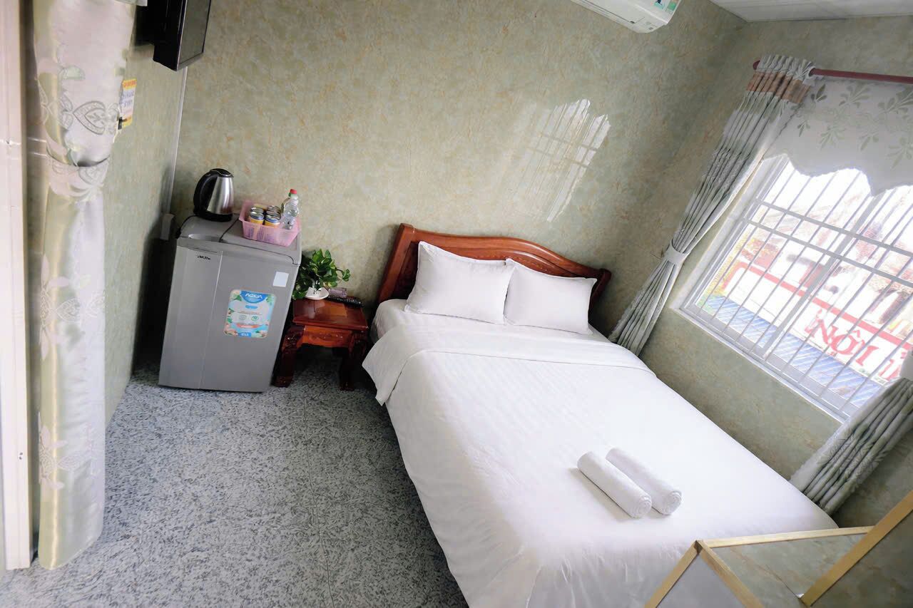 A single bed with white linens, a small bedside table with a kettle and a plant, a refrigerator, and a window with curtains.