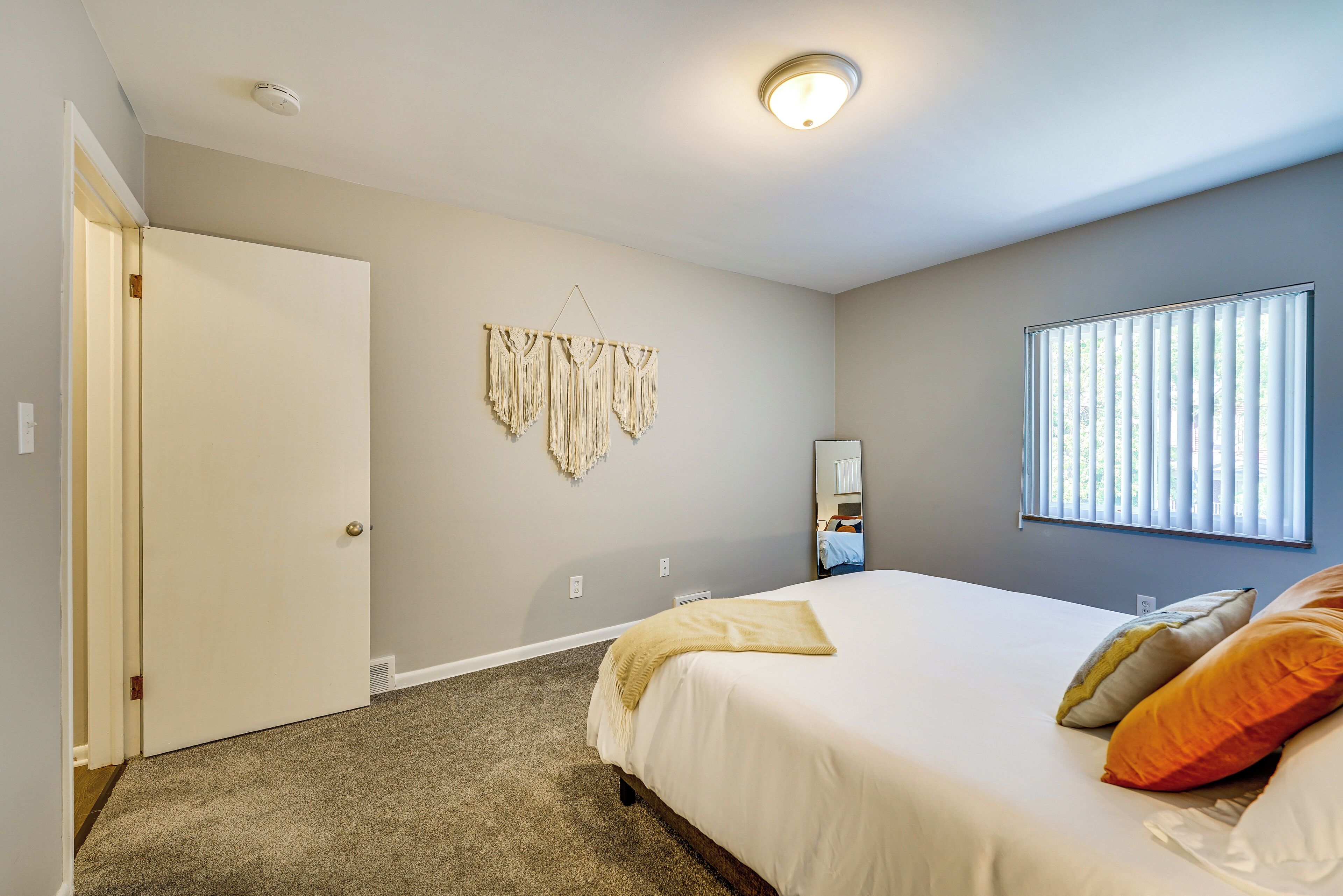 Una camera da letto con un letto, una porta, una decorazione in macramè appesa al muro, una finestra con tende e una lampada a soffitto.