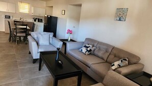 Een moderne woonkamer met een grijze bank, een zwarte salontafel, en een eethoek met een witte tafel en stoelen.