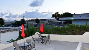 Een terras buiten met tafels en stoelen, rode parasols, en een uitzicht op een parkeerplaats en gebouwen op de achtergrond.