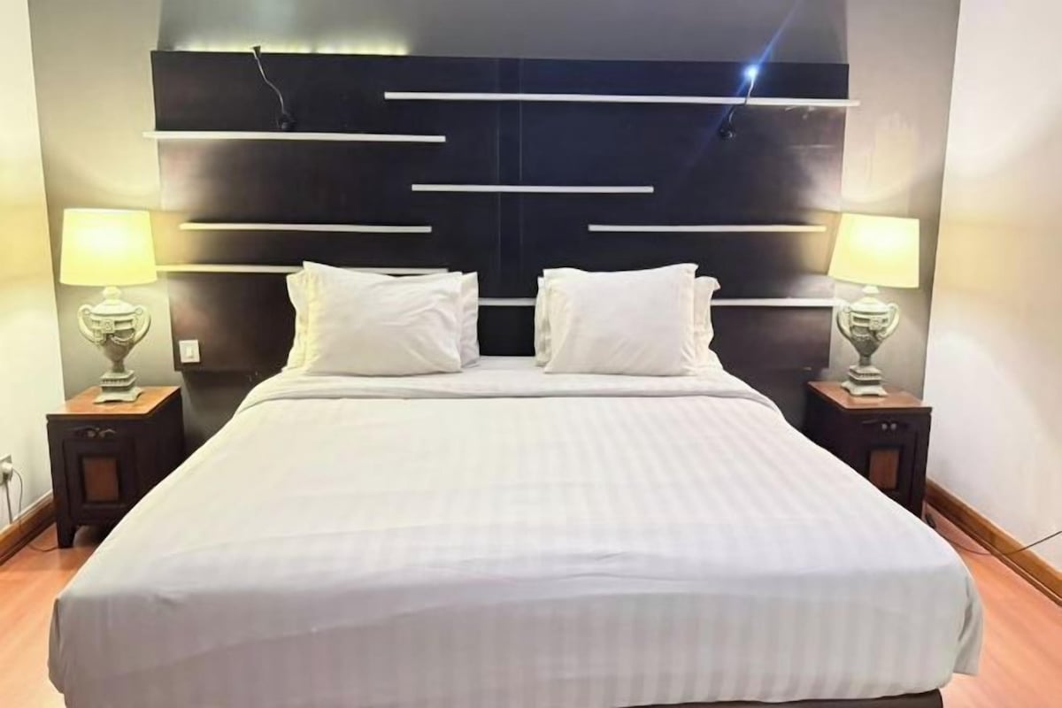 Una habitación de hotel moderna con una cama grande, dos mesitas de noche con lámparas y un cabecero oscuro con líneas horizontales.