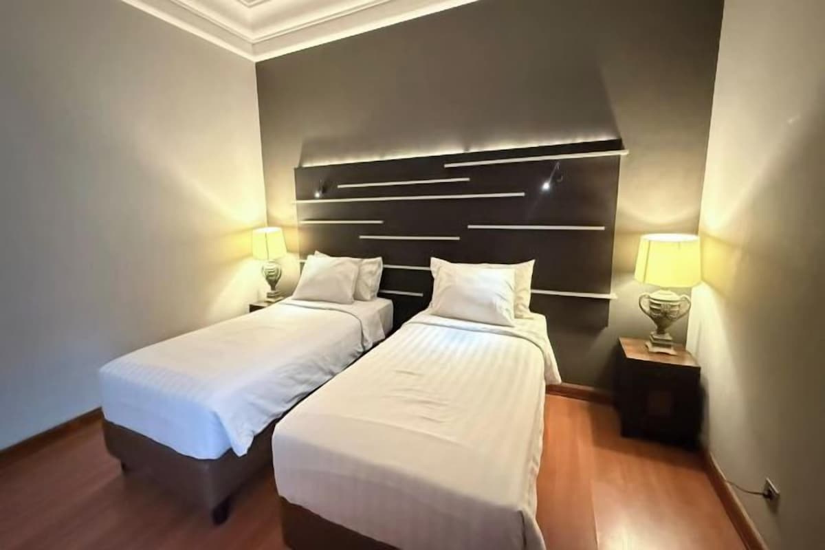 Habitación doble con dos camas, una moderna cabecera, lámparas de noche y una mesita con lámpara.