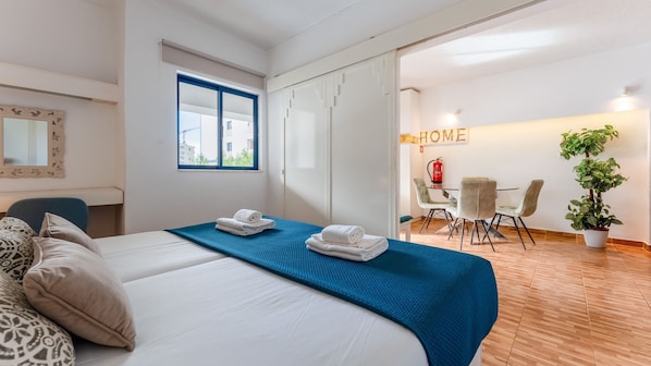 Quarto de hotel com cama, escrivaninha, cadeira, mesa de jantar com cadeiras, planta em vaso e um letreiro com a palavra 'HOME'.