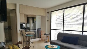 A modern living room with a grey sofa, teal chairs, and a small round table. A kitchen with a fridge and a dining area is visible in the background. - Modern apartment located in a resort style condo, pools, jacuzzi, soccer field,. (La Tebaida)