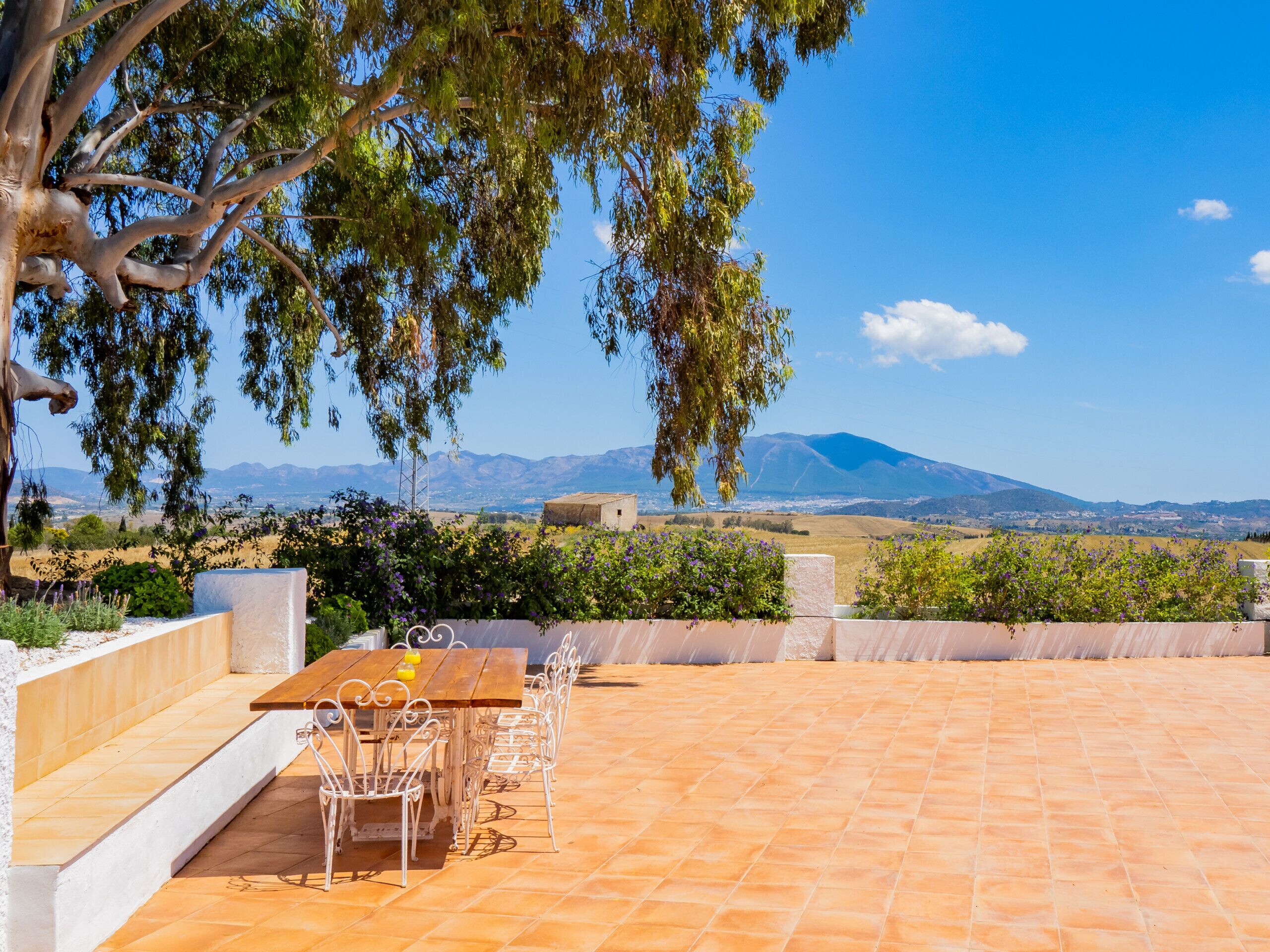 Une terrasse extérieure avec une table et des chaises en bois, entourée d’un sol en pierre et offrant une vue sur des montagnes lointaines.