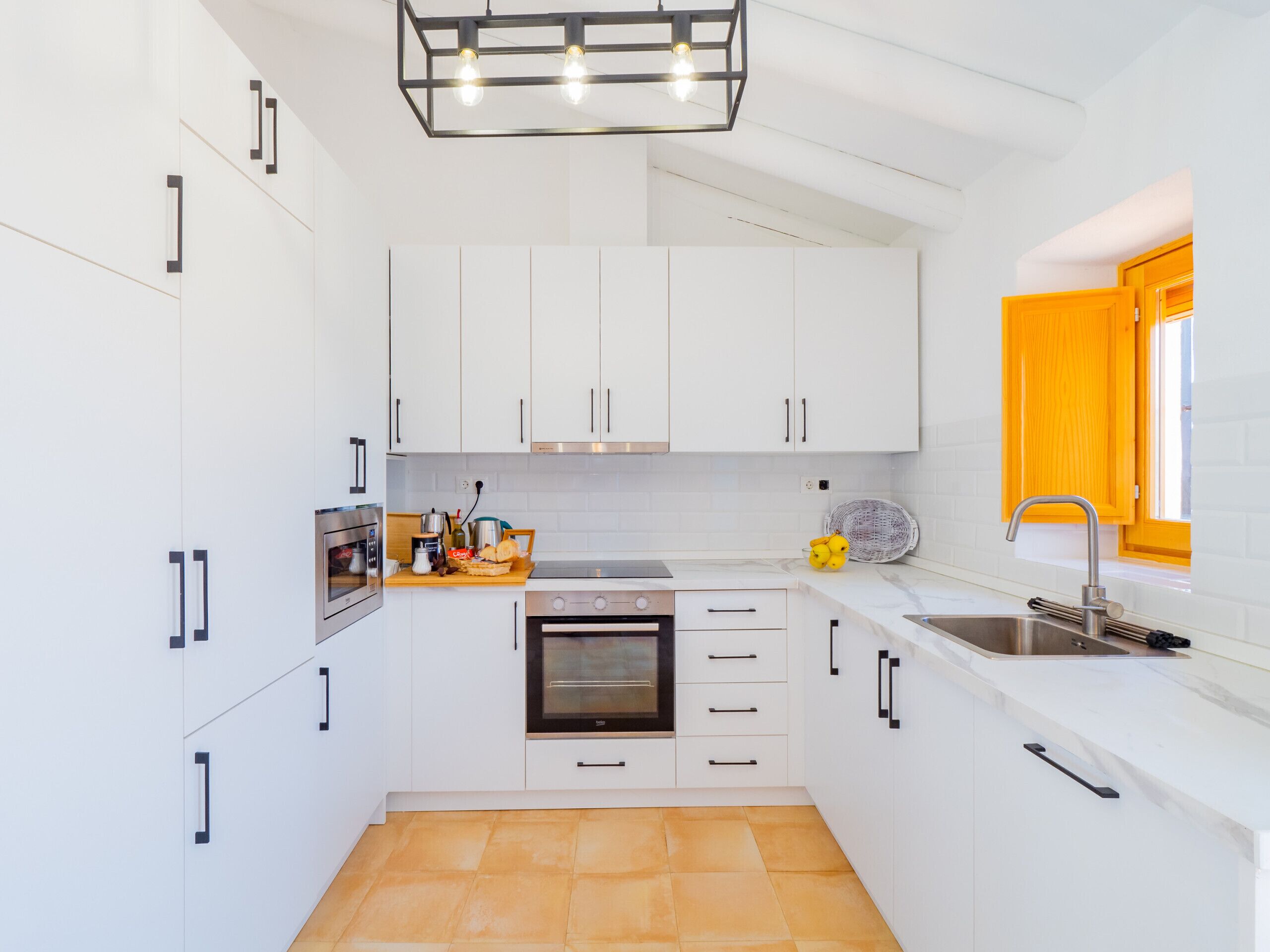 A compact kitchen with white cabinets, a built-in oven, a sink, and a window with a yellow frame.