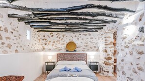 Un dormitorio con una cama grande, dos mesitas de noche, una silla y una pequeña mesa. La habitación tiene paredes de piedra y techo de madera.