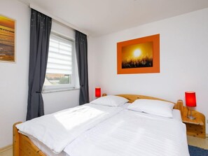 Uma cama bem arrumada com roupa branca, uma janela com cortinas, um abajur vermelho e um quadro na parede.