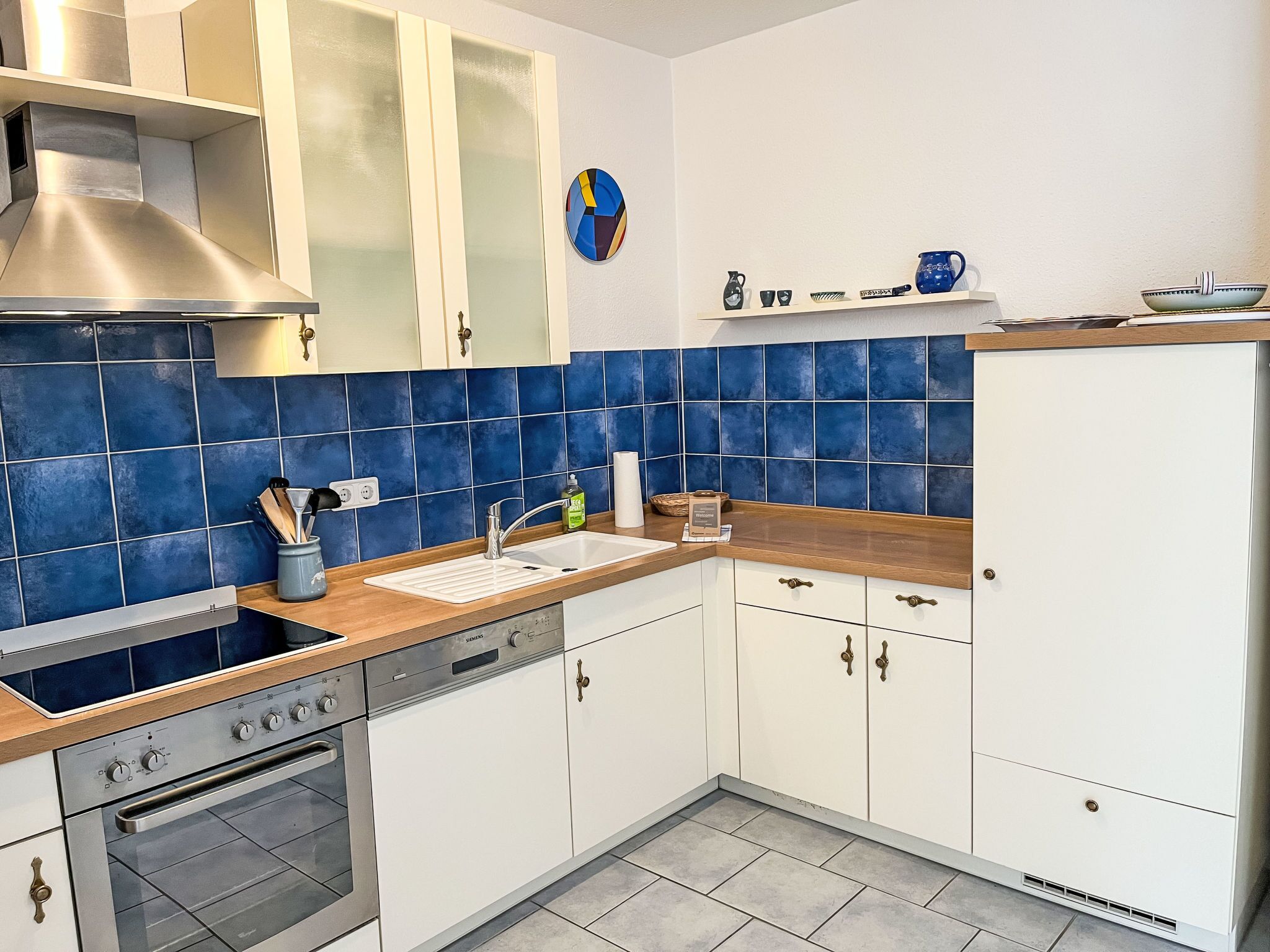 Moderni keittiö, jossa on valkoiset kaapit, ruostumattomasta teräksestä valmistettu liesi ja sinisillä laatoilla päällystetty seinä liesen takana.