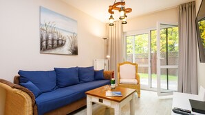 Una sala de estar con un sofá azul, una mesa de centro de madera, una silla de mimbre, un cuadro en la pared y un ventanal corredero que da a un espacio exterior.