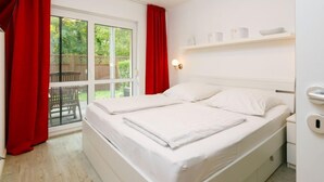 Un dormitorio con cama, una ventana con cortinas rojas, una mesita de noche blanca y un pequeño estante sobre la cama.