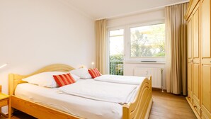 Een tweepersoonsbed met witte lakens en twee rode en oranje gestreepte kussens, een houten hoofdbord, een nachtkastje met een lamp, een raam met gordijnen en een radiator.
