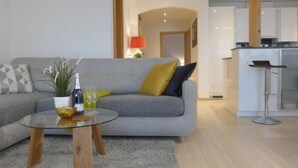 Ein modernes Wohnzimmer mit grauem Sofa, einem Glastisch und einer kleinen Pflanze. Im Hintergrund ist eine Küchenzeile erkennbar.